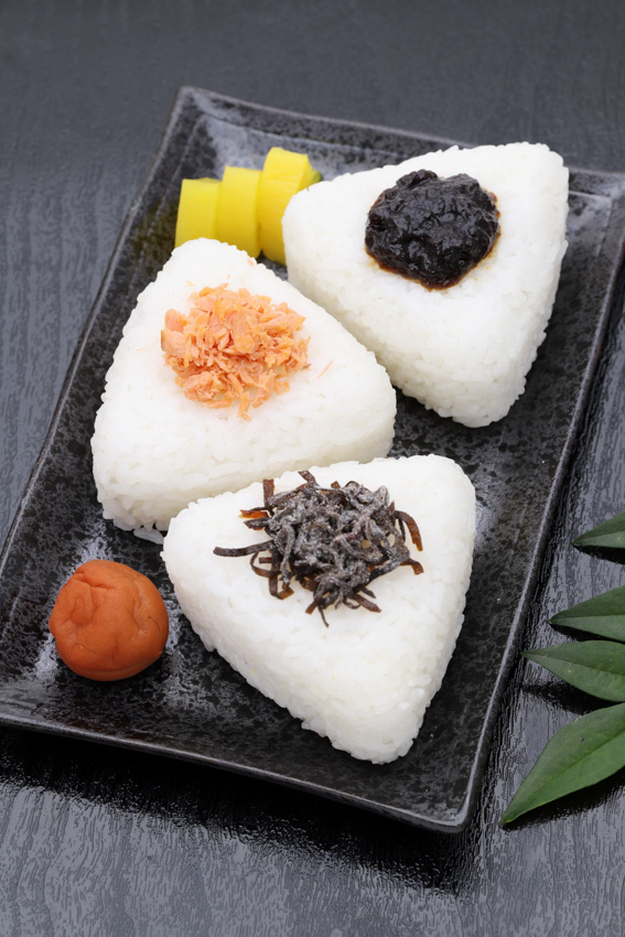 Three onigiri with fillings on a plate. Konbu, salmon, and tskudani. On the plate is also one umbeboshi and some pickled vegetables.