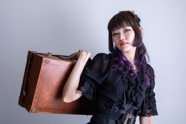 Girl with dark purple hair in a gothic type black lolita fashion dress holding a bag over her shoulder