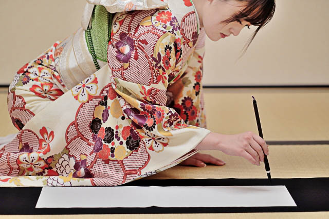 A woman in kimono in kneeling position on a tatami floor doing calligraphy.