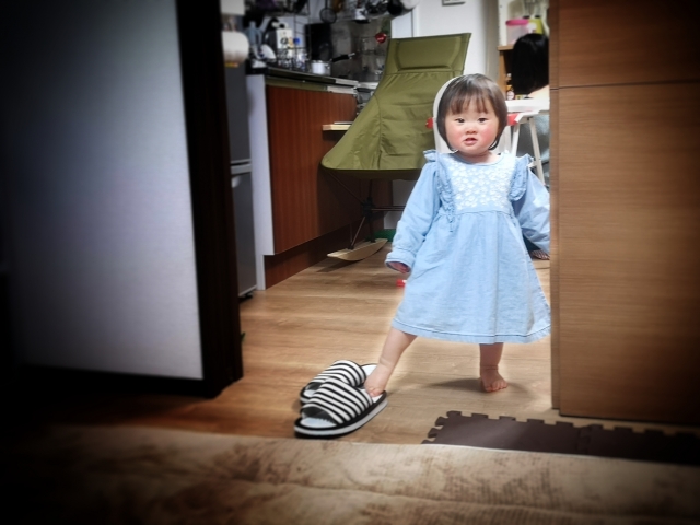 A child standing next to a door touching with the feet a pair of slippers