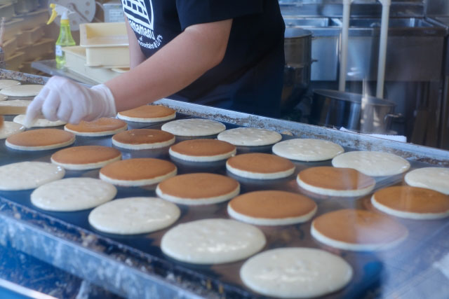 A person making Dorayaki in a shop on a big grill with dozens of Dorayaki spread out in rows.