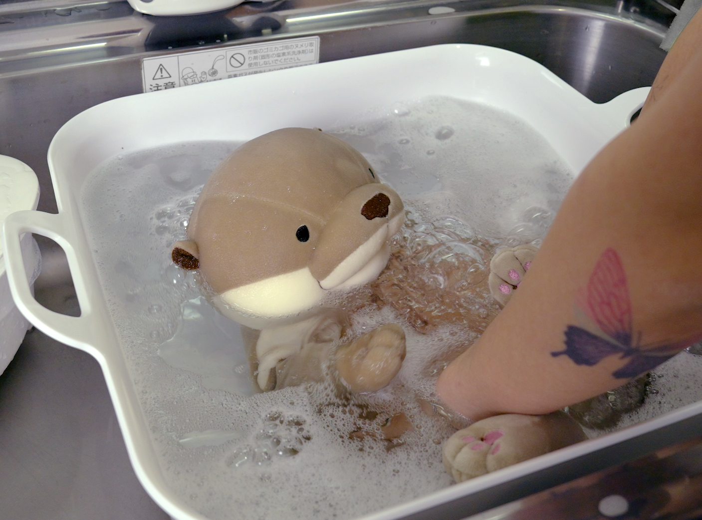 Gray stuffed otter washed in soapy water in white pail in sink