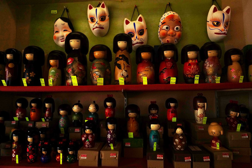 Three rows of about two dozen dolls in a doll shop with masks hanging on the wall behind