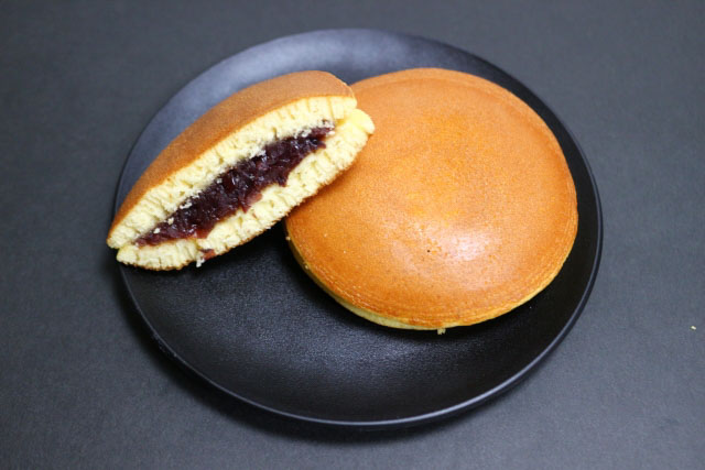 One Dorayaki cut in half to reveal the filling and one whole Dorayaki under it on a black plate.