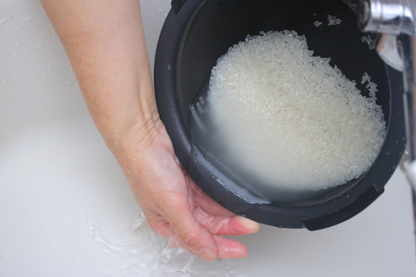 A pot of raw rice being rinsed with water from a tap.