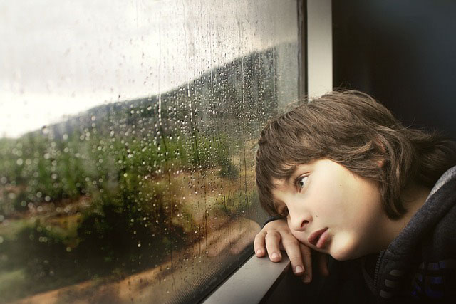 A girl resting her head on the inside of a window looking out at the rainy weather with a sad face