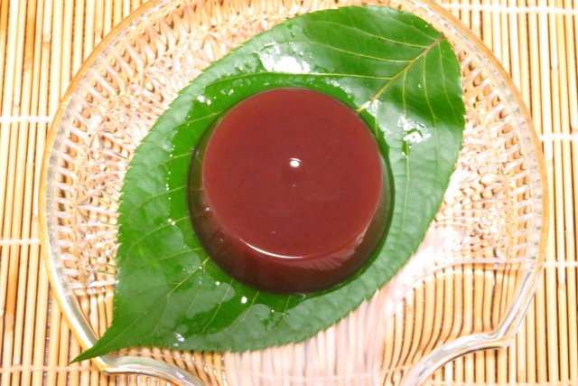 A round piece of brown yokan placed on a green leaf on top of a transparent plate on a bamboo mat