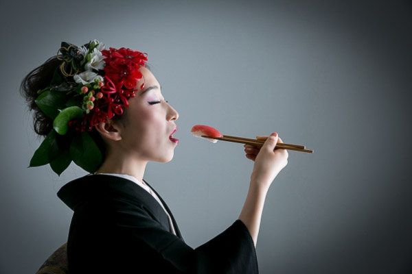 A woman wearing kimono, hair ornaments and white face make up and using chopsticks to eat a piece of sushi