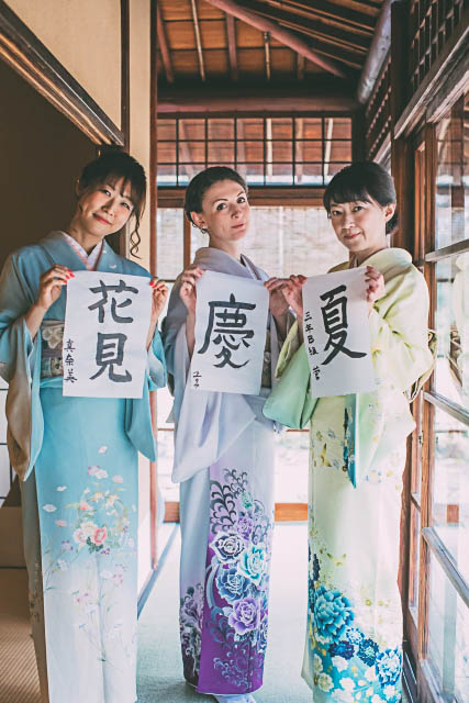 Three women in kimono standing inside a traditional style Japanese house holding a paper each with kanji characters.