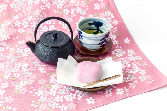 A Japanese tea set and a pink daifuku on a cherry blossom themed table cloth