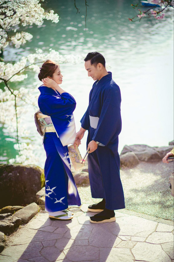 Woman and man wearing yukata standing in front of a pond with cherry trees nearby.