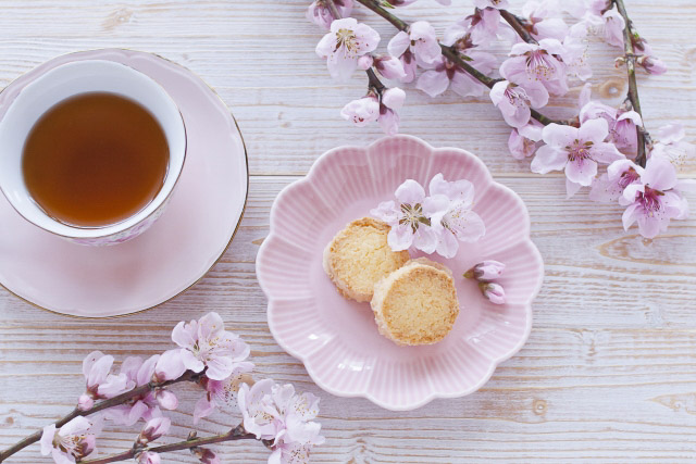 Cookies on a pink cherry blossom shaped plate with a cup of tea next to it on a light colored wooden table with cherry blossoms