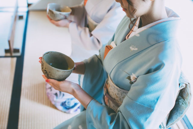 A woman in kimono sitting on a tatami floor holding a green tea cup and participating in a tea cermony.