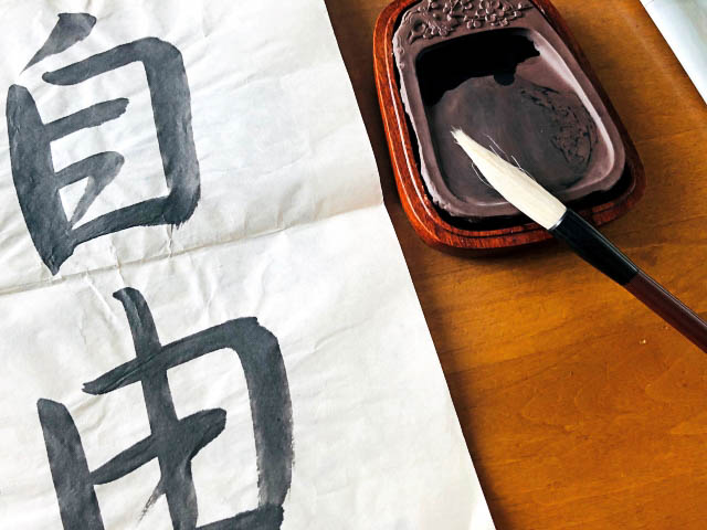 A calligraphy brush resting on an inkstone with ink next to a paper with kanji characters written on it.