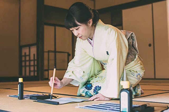 A woman in kimono in kneeling position on a tatami mat doing calligraphy.