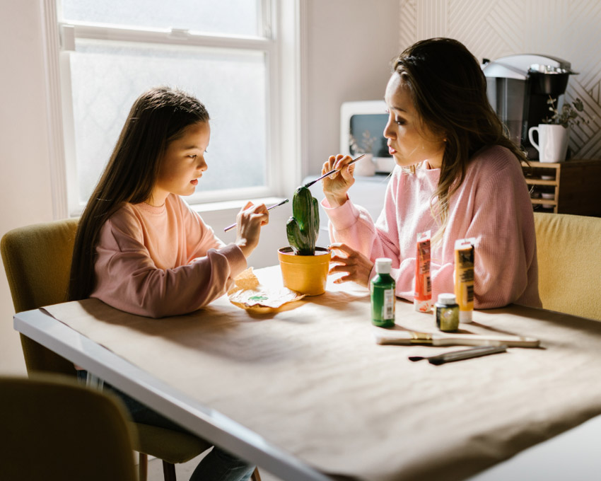 A woman and an elementary school age child at a kitchen table painting a model of a cactus