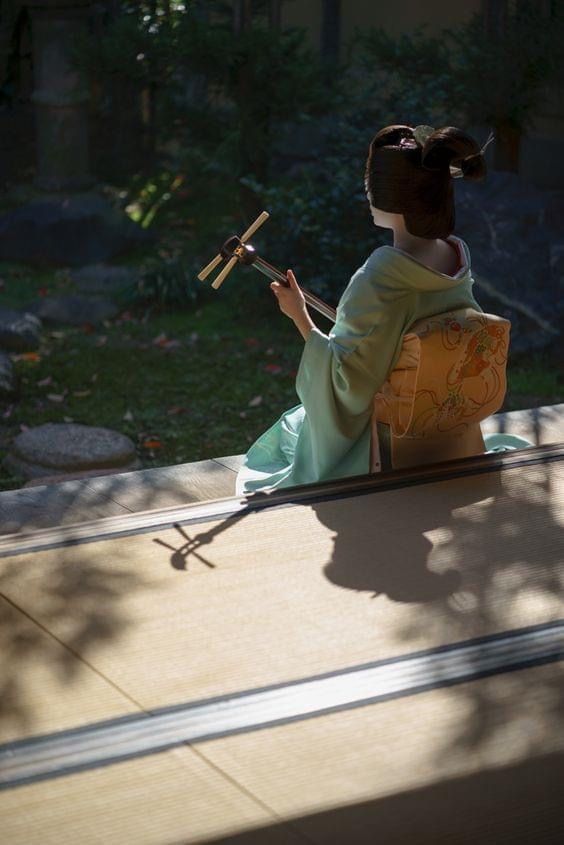 An image of someone dressed as a Maiko playing the Japanese instrument called Shamisen near a garden