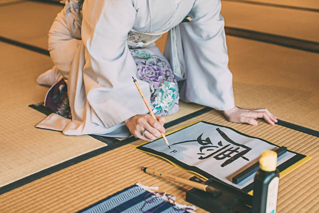 A person wearing kimono sitting in a kneeled position, writing a kanji character with a brush.